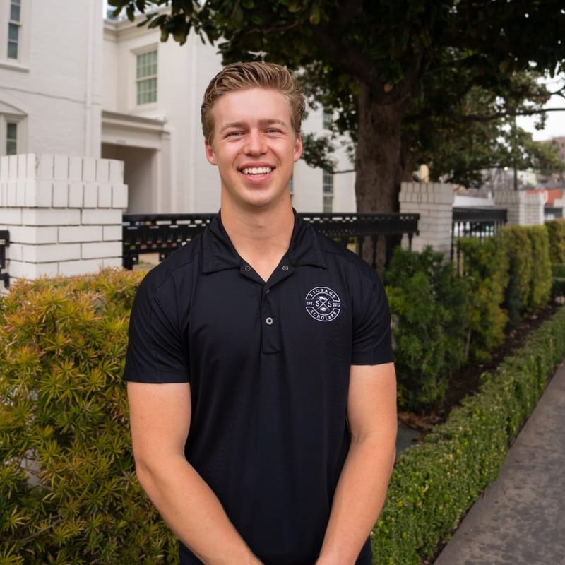Matt Gronberg smiling outdoors in a black Storage Scholars polo shirt, standing in front of hedges and a white building