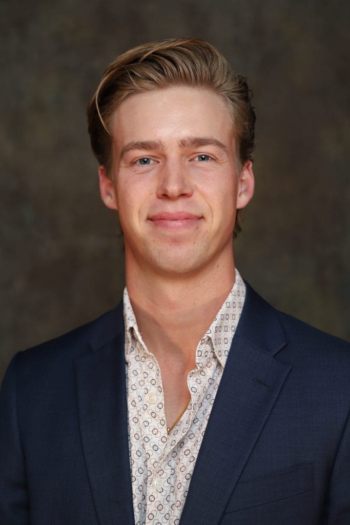 Matt Gronberg headshot, wearing a navy blazer and patterned open-collar shirt against a neutral brown background