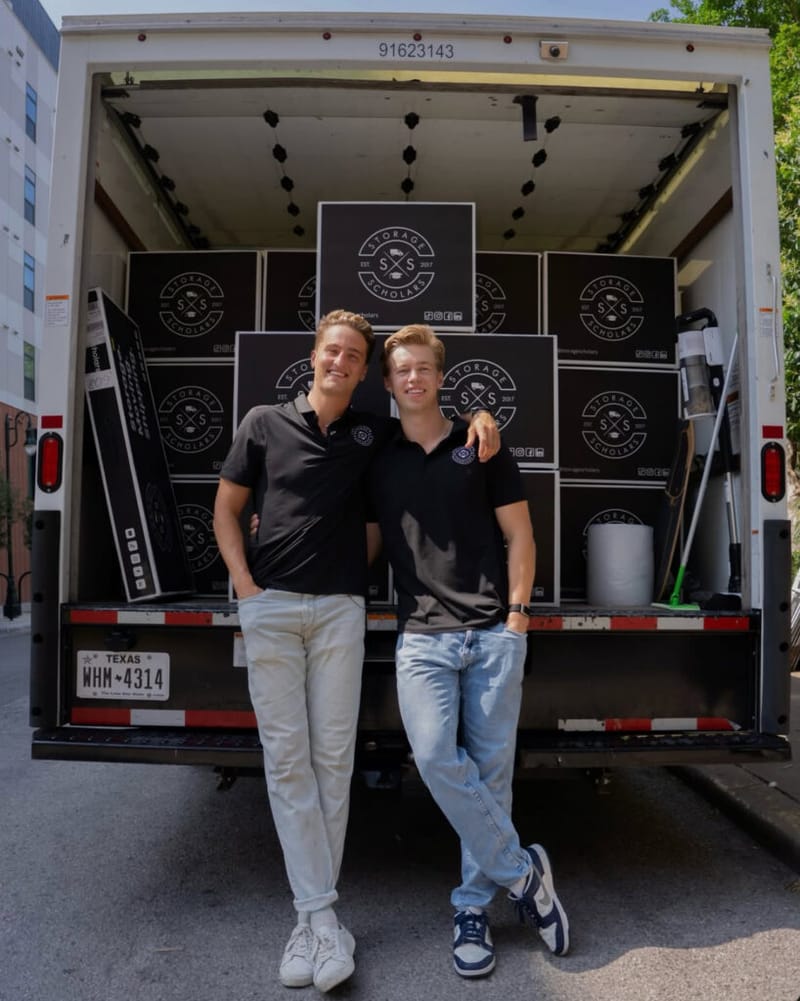 Two Storage Scholars co-founders in black polo shirts standing in front of an open moving truck packed with branded boxes after a spring move-out
